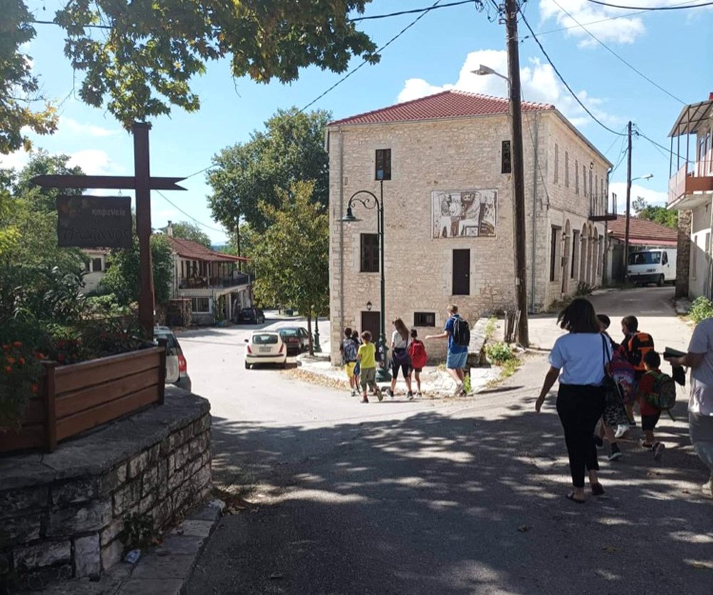 Ομάδα ανθρώπων περπατά στην πλατεία της Πωγωνιανής μπροστά από πέτρινο κτίριο